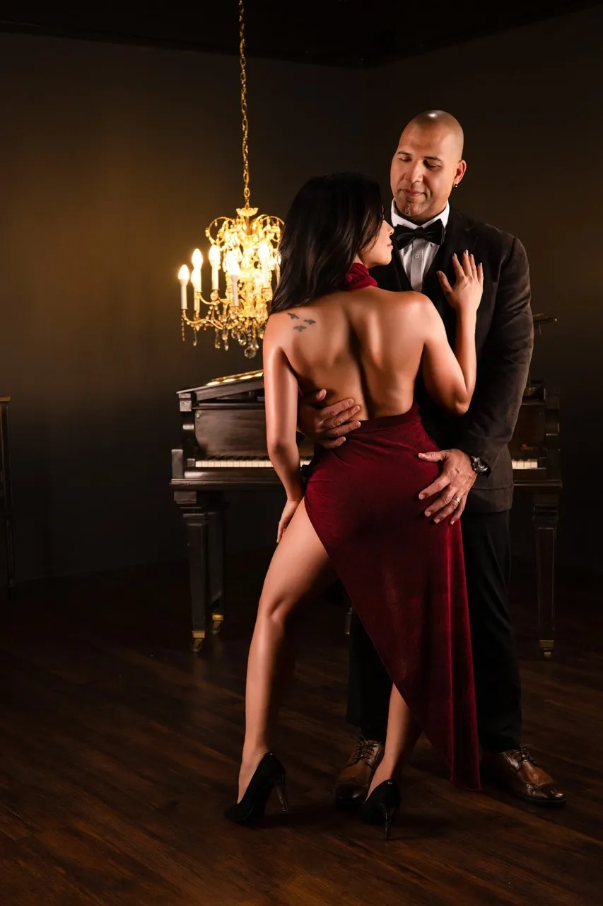 Romantic couples boudoir portrait featuring a woman in a backless burgundy dress with a high slit, embracing her partner in a dark suit beside a grand piano. Warm chandelier lighting emphasizes her exposed back, body curves, and the couple's intimate connection.