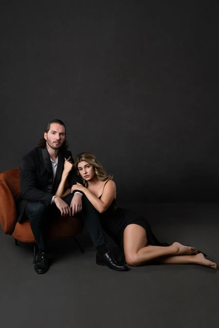 Sophisticated couples boudoir portrait featuring a man in a tailored black suit seated in a velvet chair while his partner, wearing a black dress, rests beside him with her legs extended. The pose highlights elegant body lines, confident expressions, and classic fashion-inspired composition.