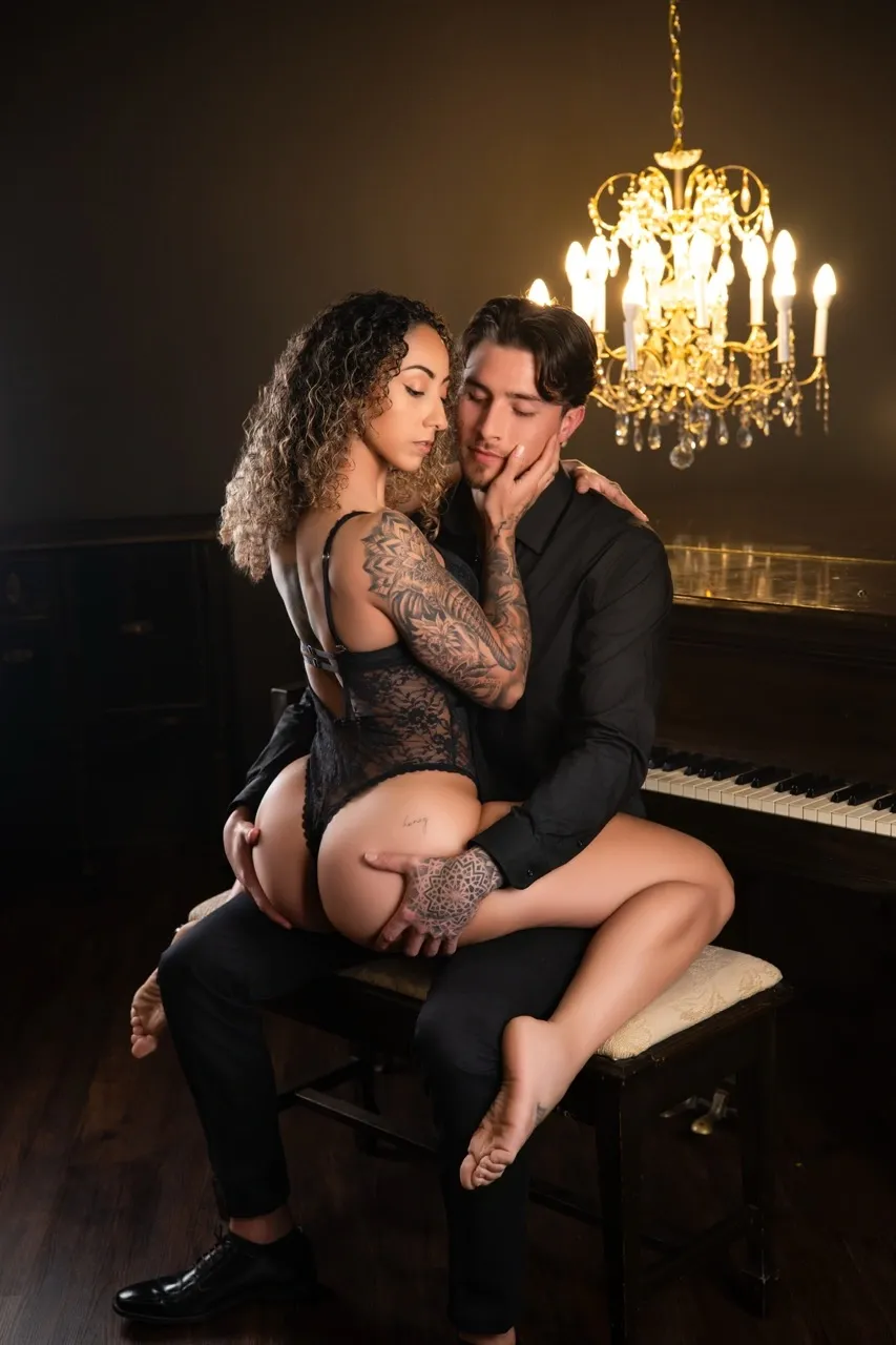 Romantic couples boudoir photography featuring a tattooed woman in black lace lingerie sitting across her partner's lap, gently touching his cheek as he holds her thighs. Their closed eyes, soft facial expressions, and close embrace communicate tenderness, trust, and emotional intimacy.