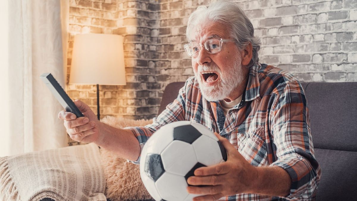 Älterer Fußballfan mit Fernbedienung und Ball jubelt auf dem Sofa vor dem Fernseher