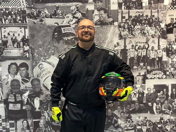 Eu, vestindo um macacão de piloto de corrida, na frente de um painel com imagens de pilotos e corridas de kart. Eu estou segurando um capacete.