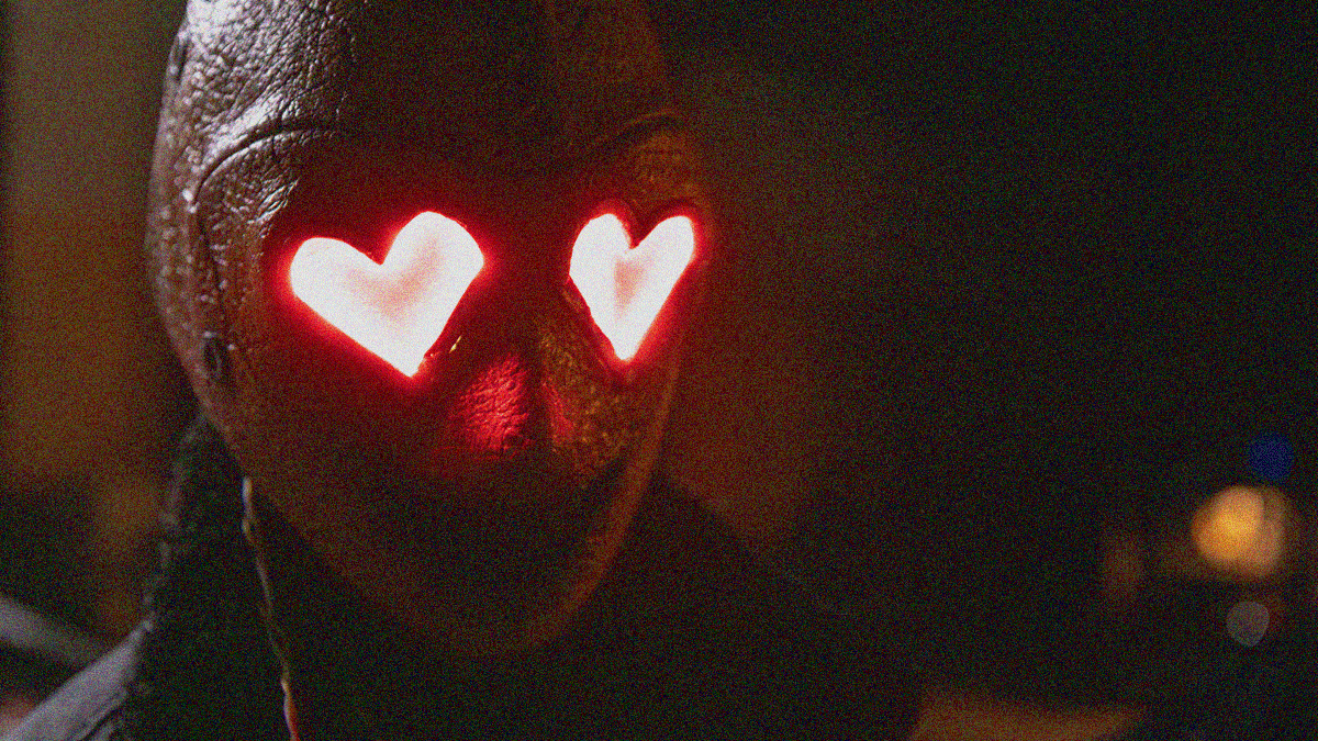 Close-up of the Heart Eyes Killer in sewn leather mask with heart-shaped glass-paned eyes glowing red.