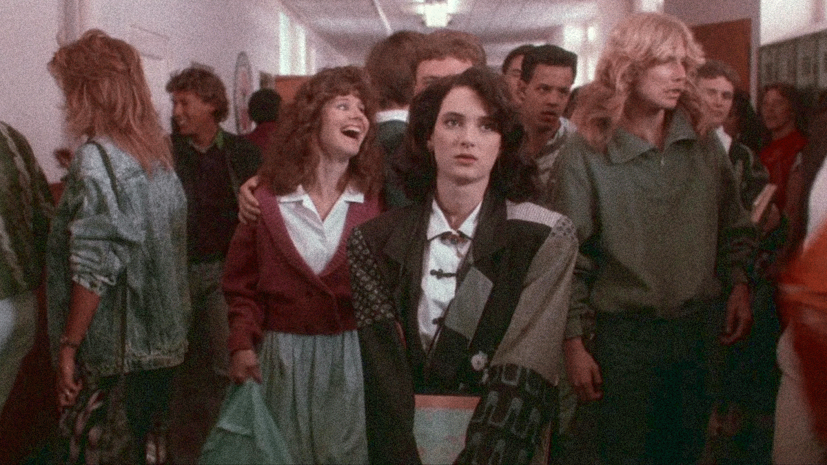 Winona Ryder walking towards the camera through a school hallway with a crowd of students behind her.