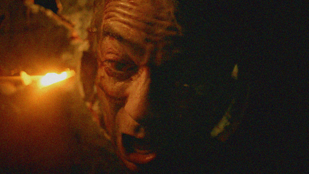 A bloodied man screams in darkness, captured from below.