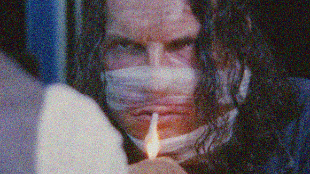 A man with gauze wrapped over his nose and chin stares menacingly as someone lights the cigarette in his mouth.