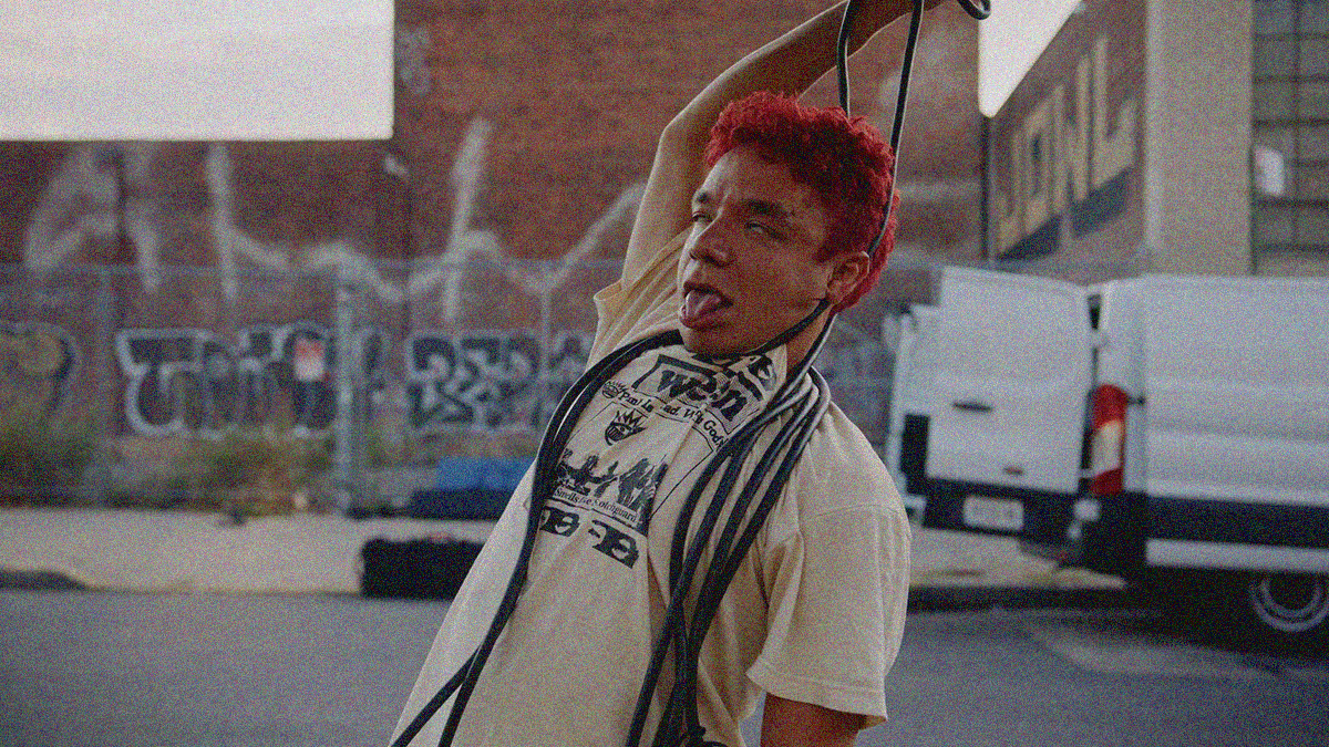 A man wrapping film equipment cords around his neck to simulate hanging himself while outside in front of a graffitied building and white van..