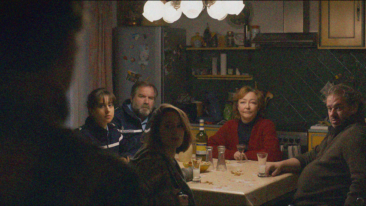 A table of people (including to police officers) all turn towards the camera to look at a man entering the room shrouded in shadow in the foreground.