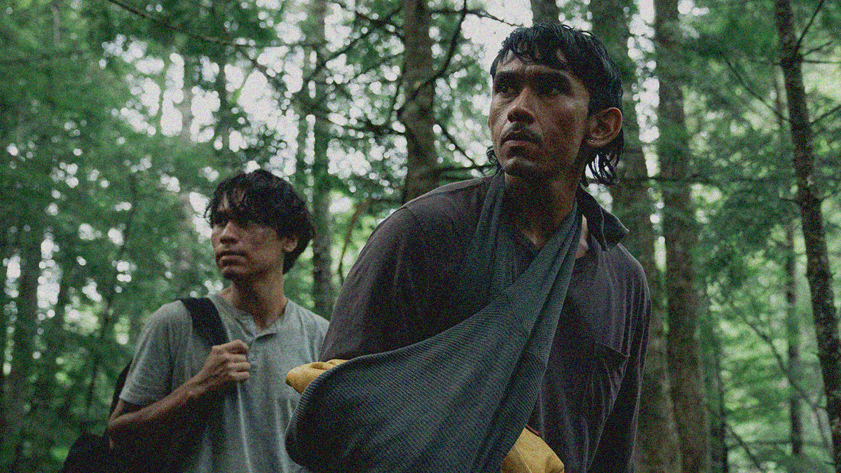 Two people stop and gaze off-screen in the forest, the one in the background holding onto a backpack and the one in the foreground with arm in a sling.