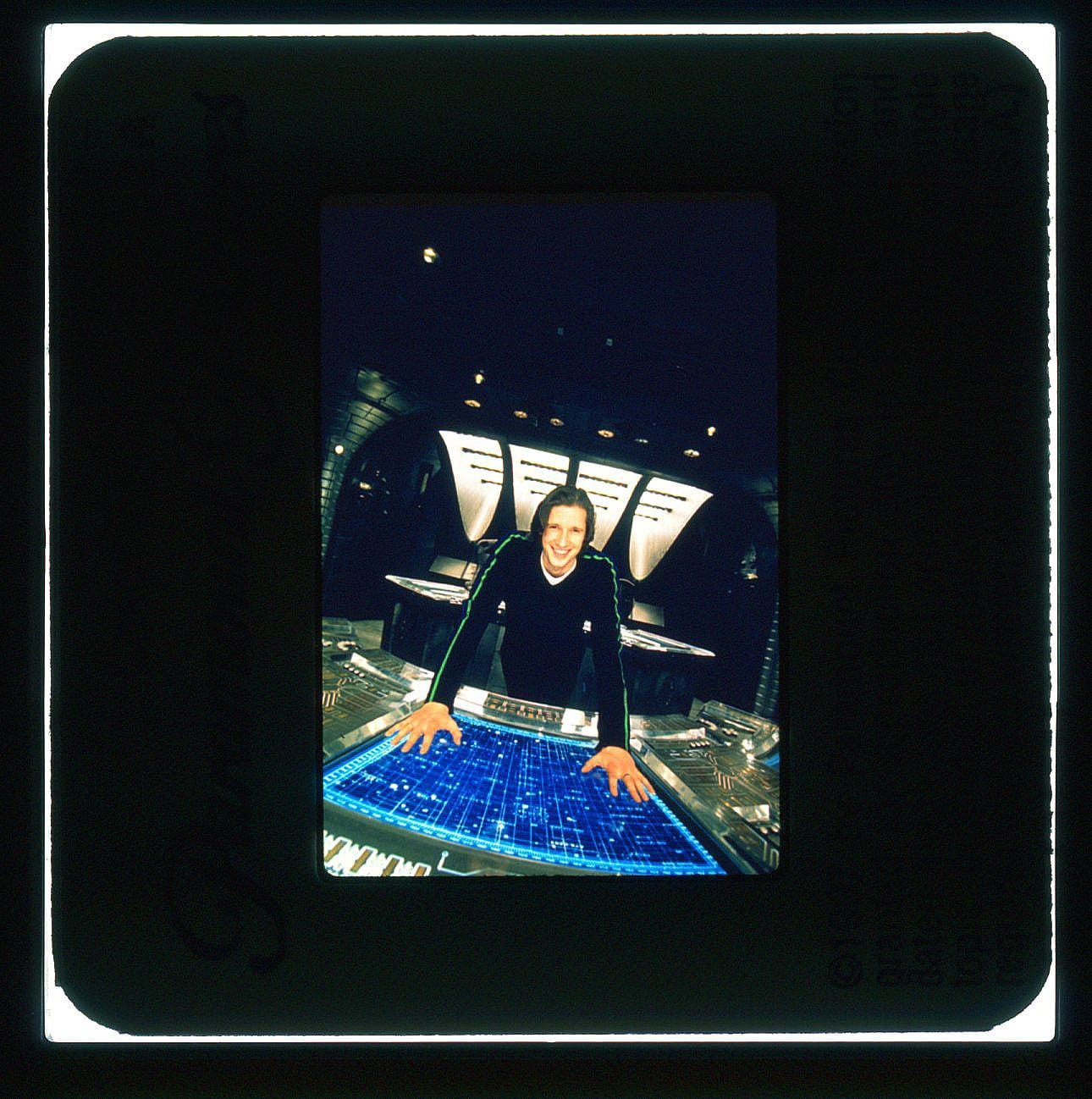 A man stands smiling with two hands planted on a spaceship's control screen below him.