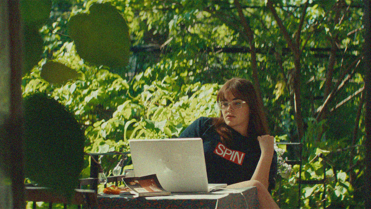 A woman leans back in her chair as she looks at the screen of her laptop outside. Green foliage fills the frame behind her.