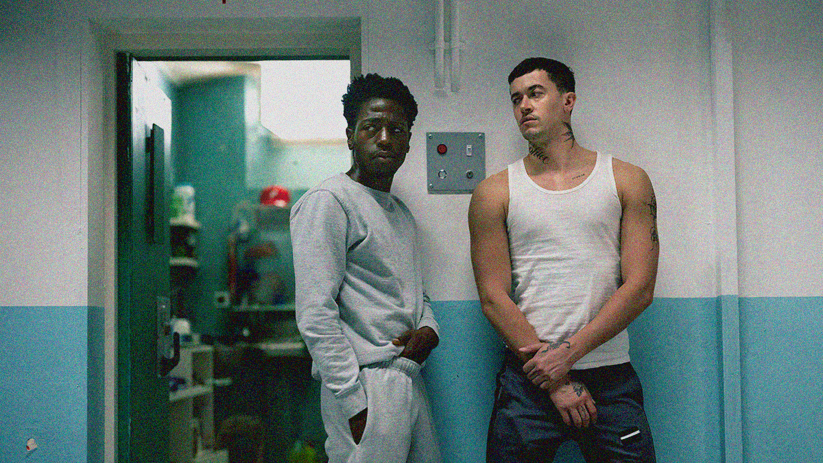 Two men stand outside the open door of a prison cell—one looks off-screen, the other looks at him.