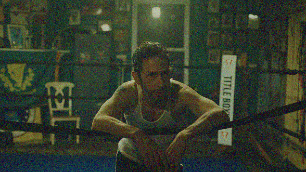 A man in a tank top leans forward on the rope of a gym boxing ring.