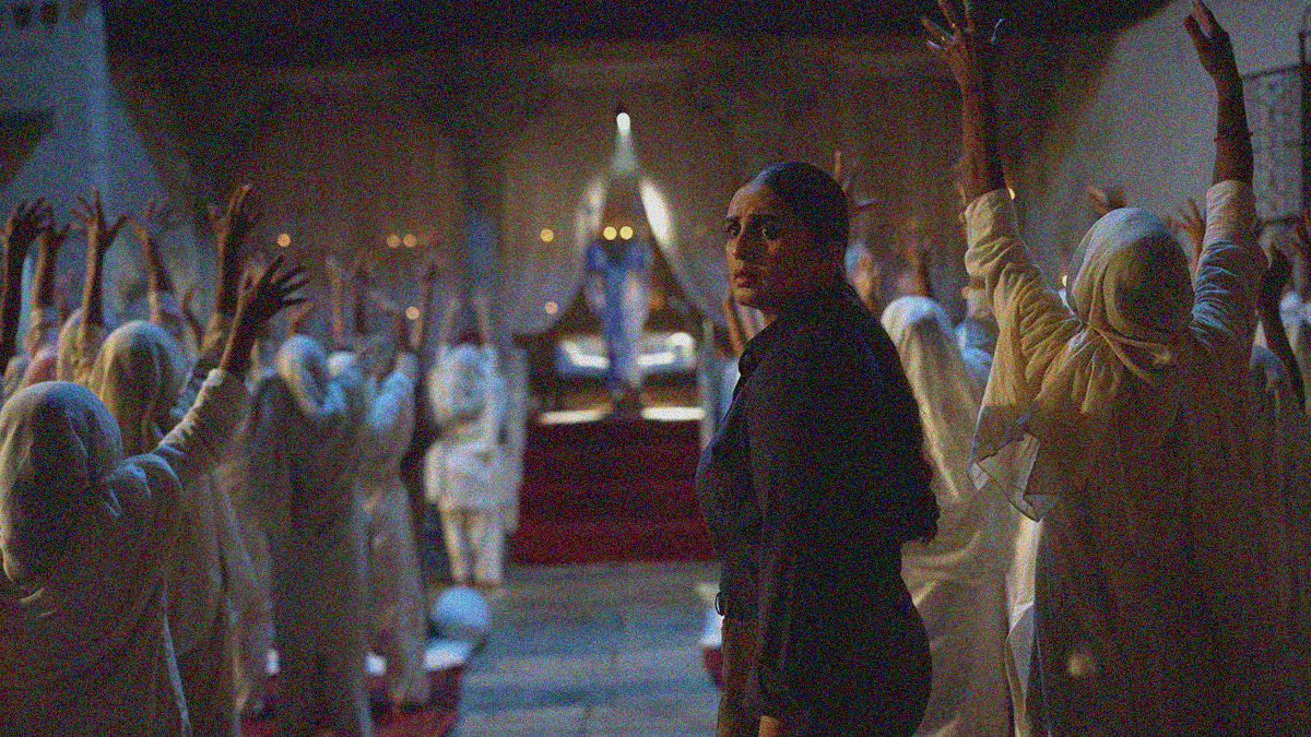 A police woman in a dark uniform stands and faces the camera as a temple full of women in white face their "Holy Father" with arms raised to the sky.