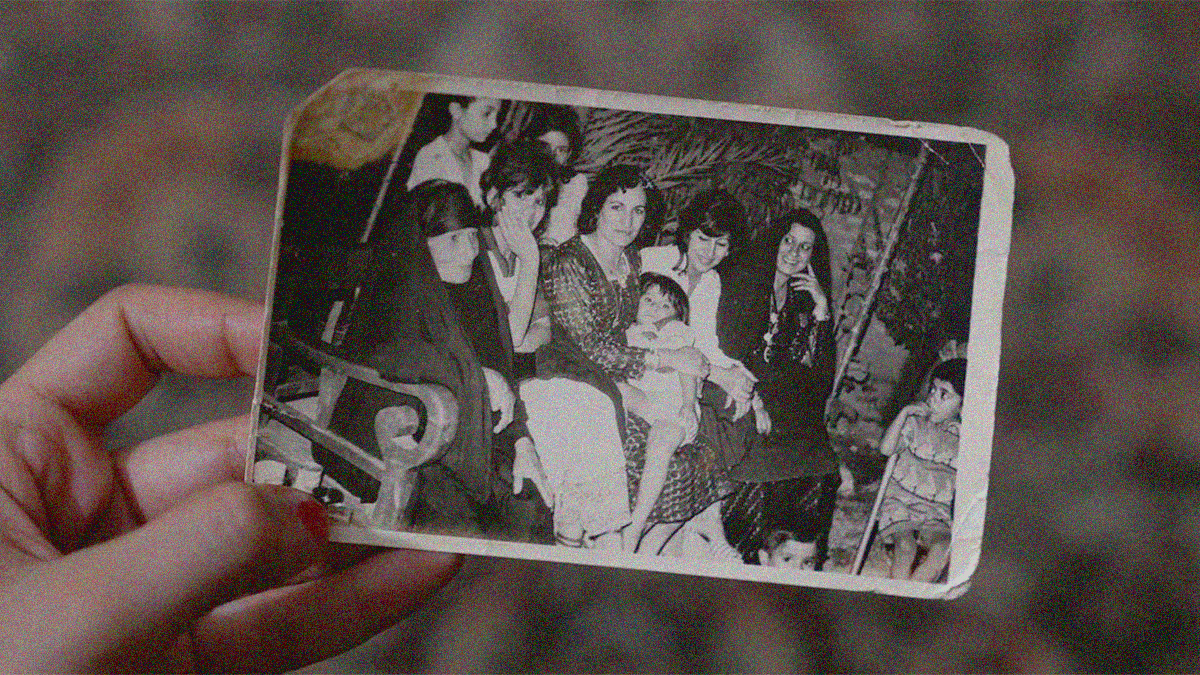 A hand holds an old photograph of a family sitting together on a long bench—five women and four children.
