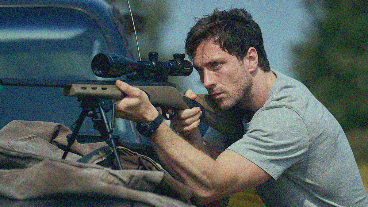 A man looks through the scope of a rifle propped up on the back of truck.