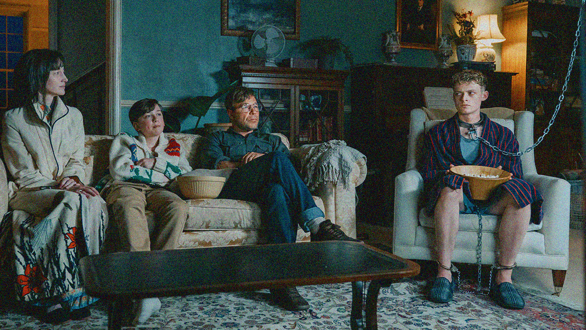 A mother, father, and son sit on a couch while another older teen sits on a chair with a collar around his neck attached to a chain going up and off-screen.