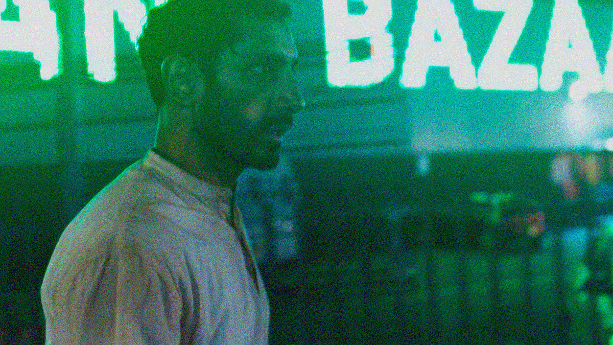 A man in profile facing right against a city street with a giant neon sign that says "bazaar".