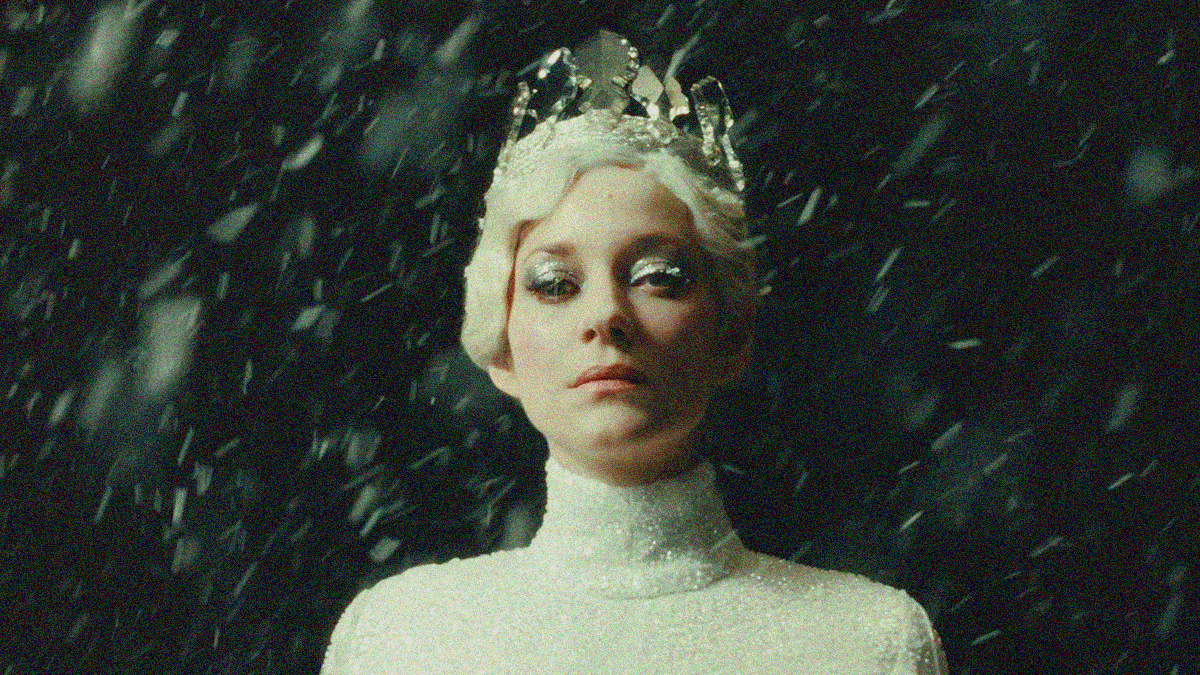 A woman from shoulders up stares at the camera. She dressed in an ornate white piece whose collar rises to her chin. She also has white hair and a crown of ice shards. It's snowing.