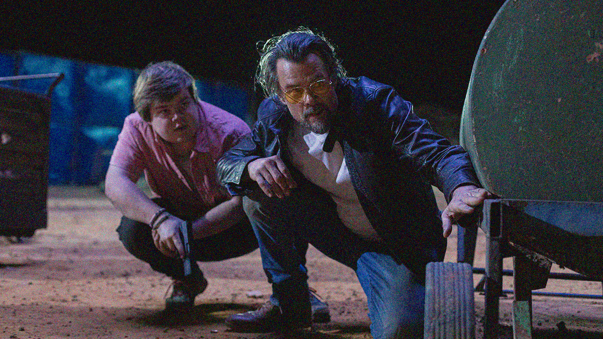 Two men crouch to peer around an obstacle. The younger one holds a gun, the older one wears glasses.