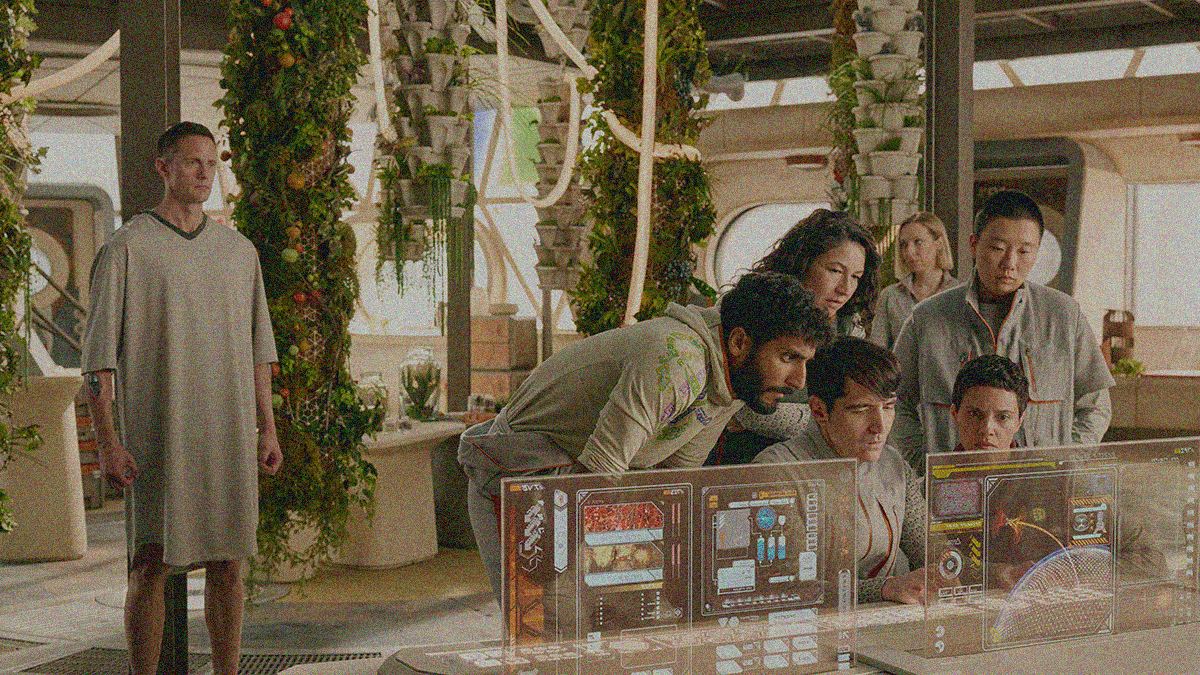 A group of people huddle around a computer screen as Murderbot stands watching from behind them.