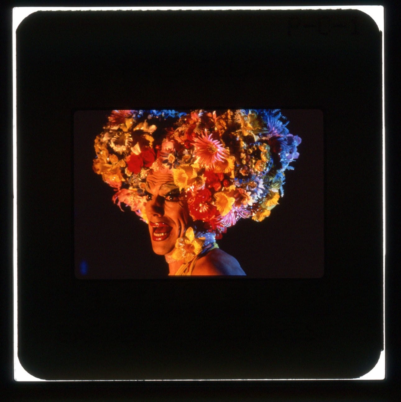 A drag queen in close-up profile with mouth open singing and a huge collection of flowers as a hat.