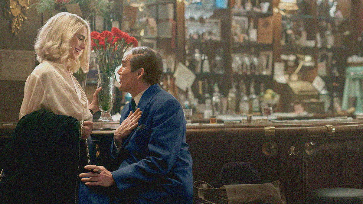 A scene at a bar with a young blonde woman standing and laughing with a vase of flowers in her hand while looking at an older balding man talking to her while sitting on a stool.