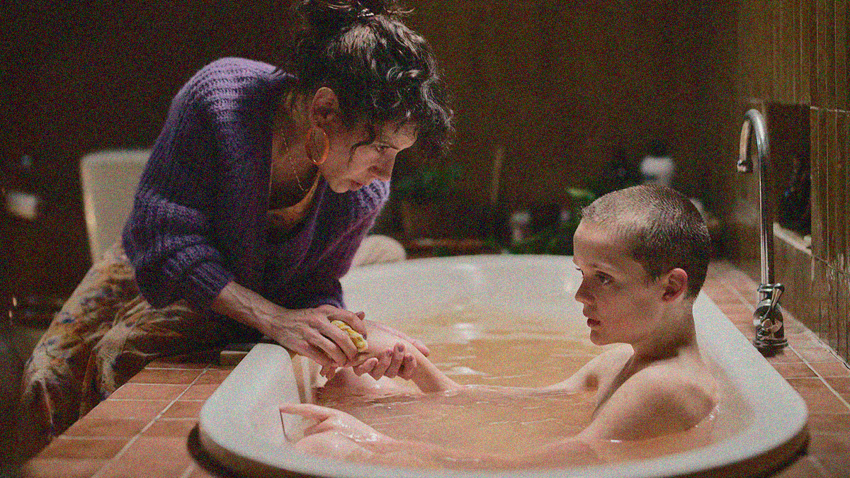 A woman leans over the edge of a bathtub as a boy sits in the water. She holds a washcloth to his hand.