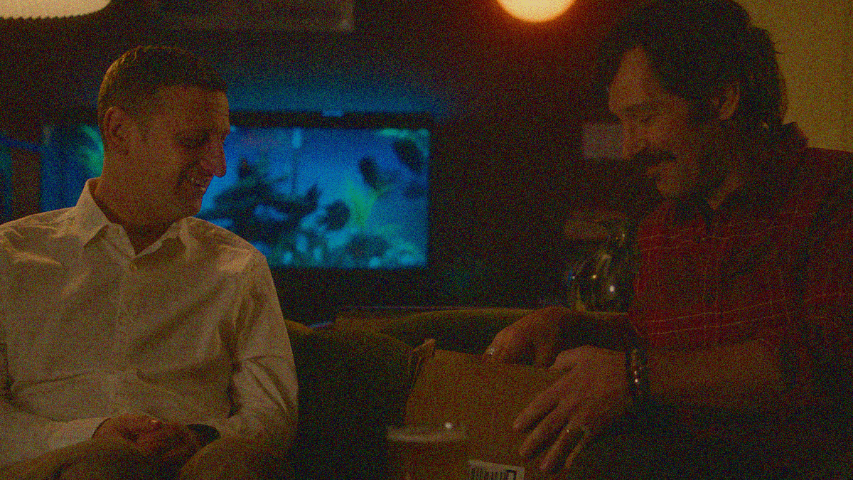 Two men sitting on a couch facing each other. One watches as the other opens a cardboard box.