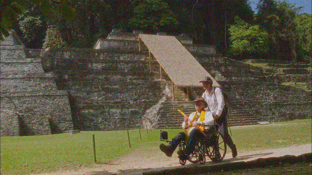 A man in a ball cap pushes an older man in a wheelchair down a path with a South American step pyramid in the near background.