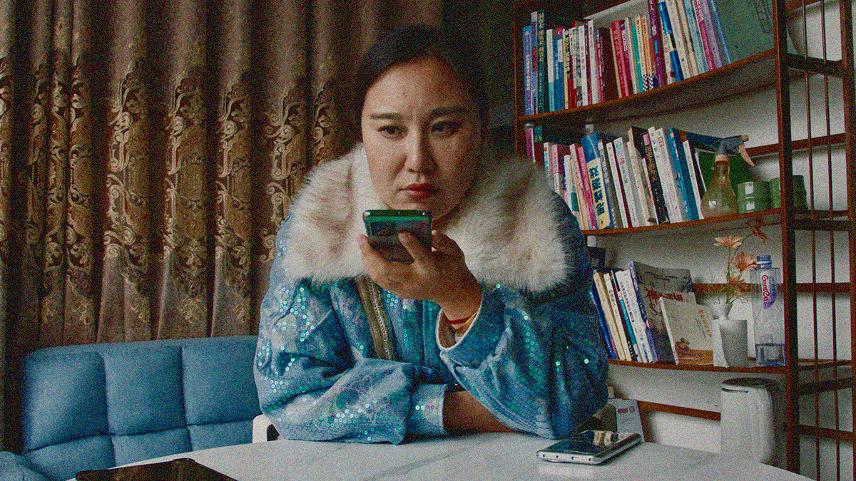 A woman sits at a table with curtains above right shoulder and bookshelf above right. She holds her cellphone up to her mouth.