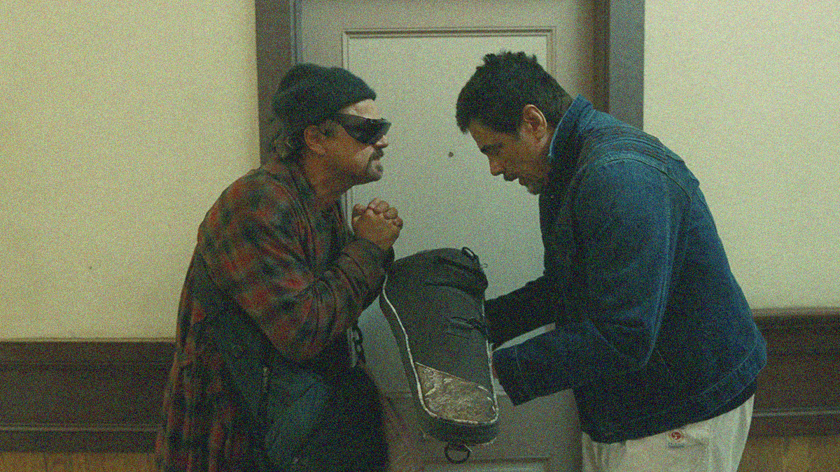 A man in sunglasses has hands clasped in a begging position opposite another man with arms outstretched holding a gun bag.