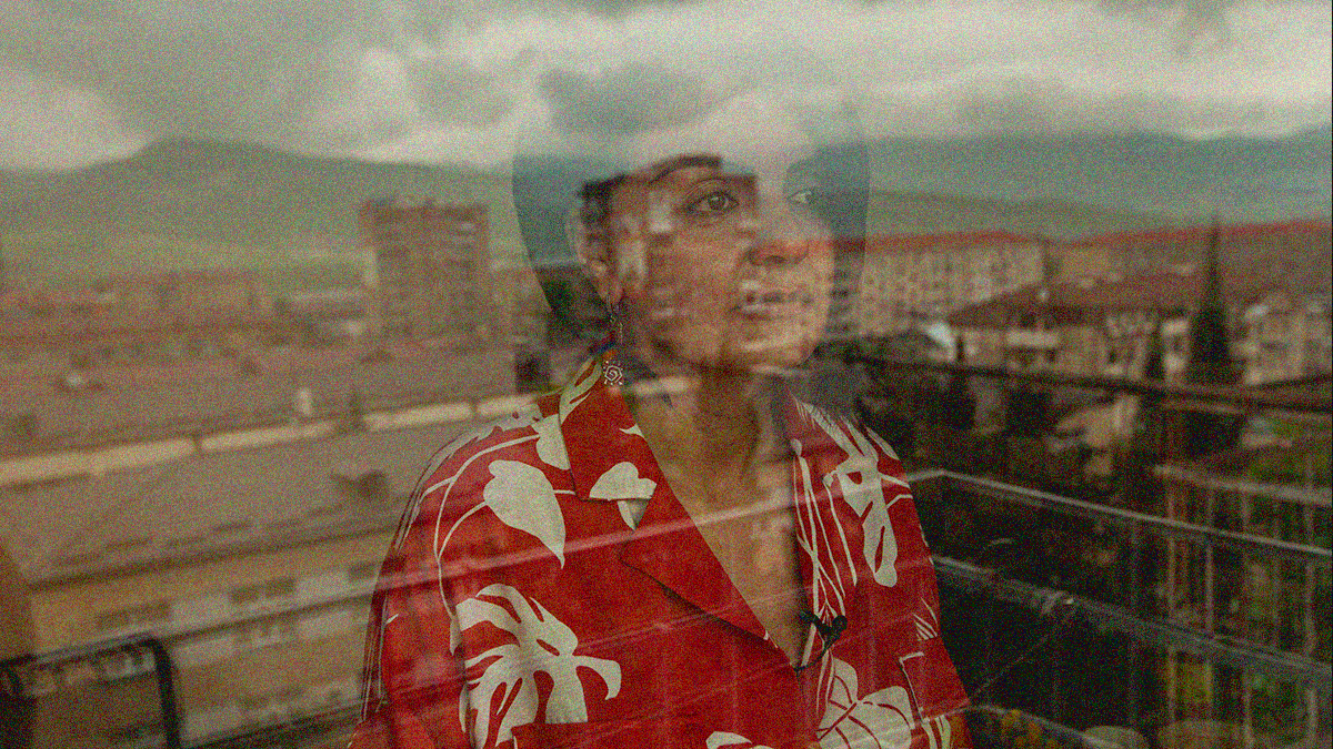 A woman in a red blouse with white silhouettes of leaves stares out a window. We see her through the glare and reflection of the world she's looking at.