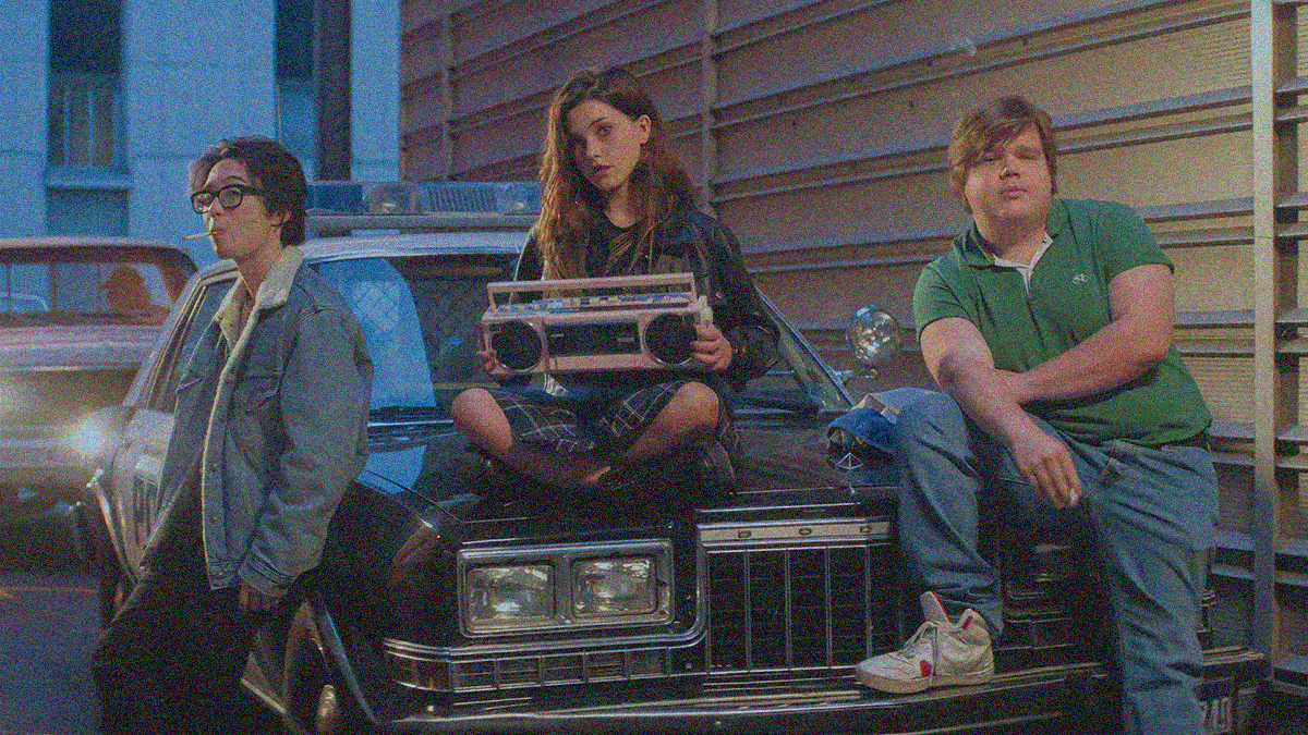 Three teens on a police car. One on left leans against the hood with cigarette in mouth. One in middle sits on hood with boombox in her lap. One on the right leans with one foot on the bumper.