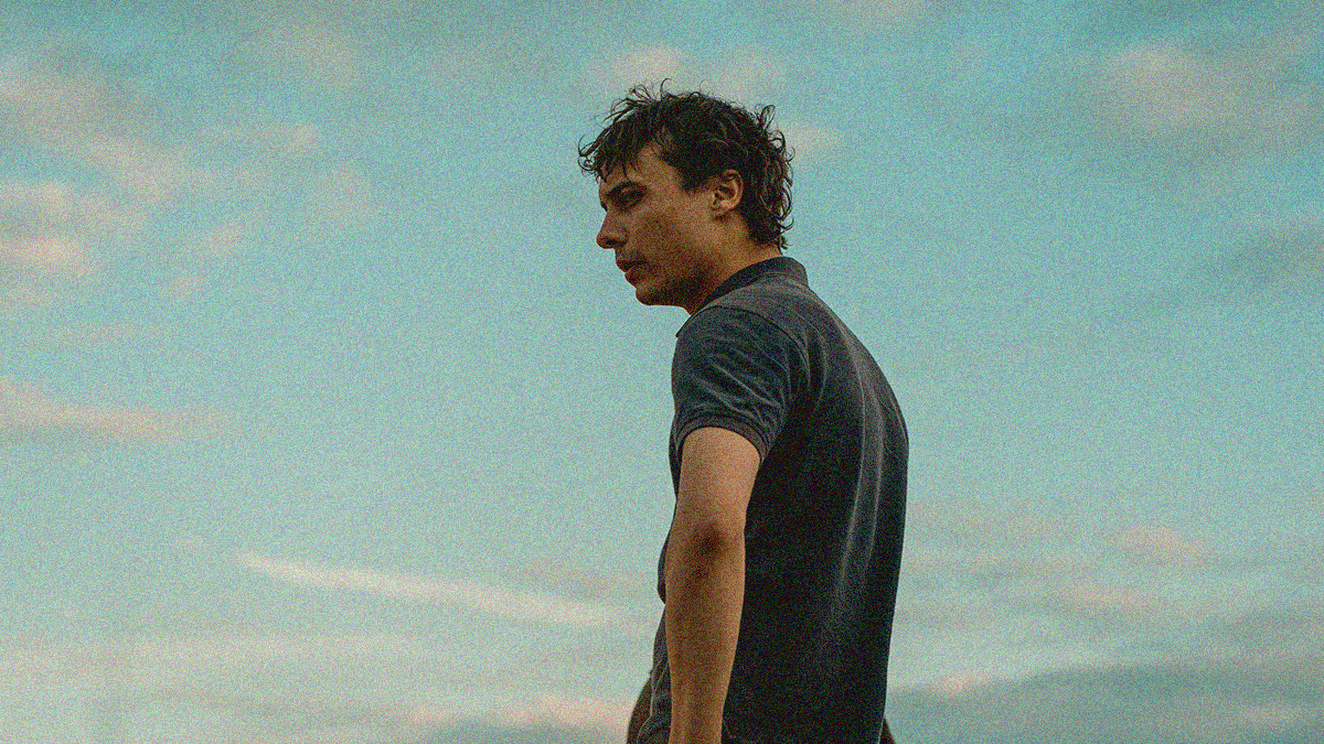 A man in profile from waist up with hands on hips is at center looking left as the sky fills the rest of the frame.