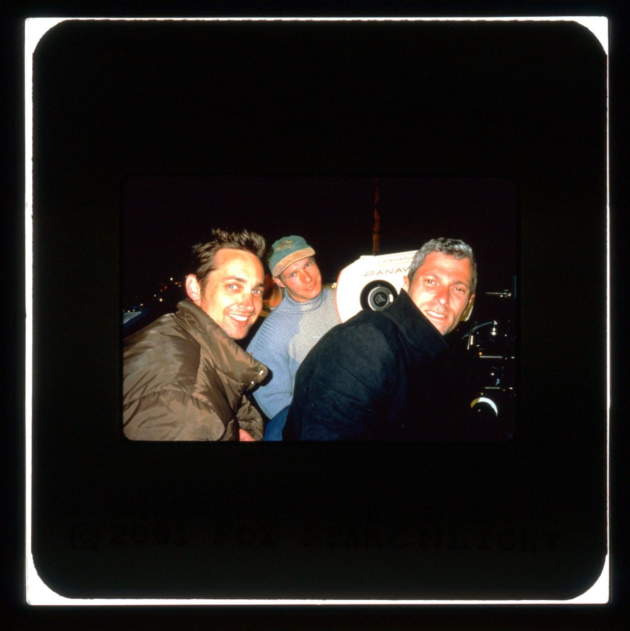 Two men in big winter coats look at the camera. A third man in a ball cap is seen between them with the film camera.