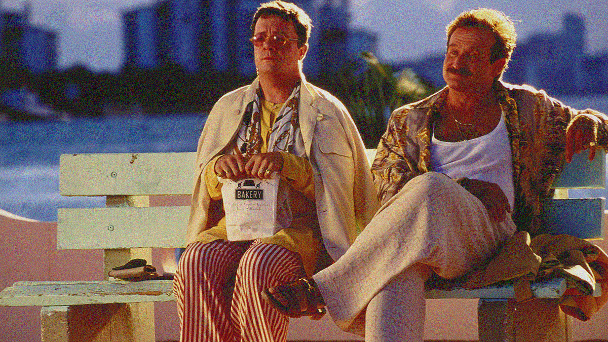 Two men sit next to each other on a park bench. Man on left sits with hand on a bakery bag in lap. Man on right has one arm on seat back and legs crossed.