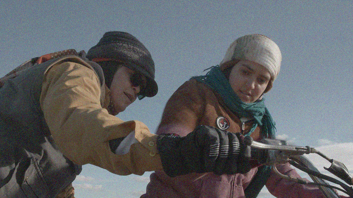 A woman on the left helps a teen woman at right learn to start a motorcycle. The latter sits on the vehicle, the former holds the handle.