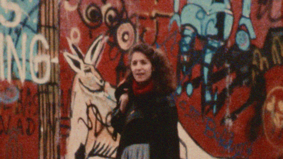 A grainy shot of a woman standing in front of a graffitied wall.