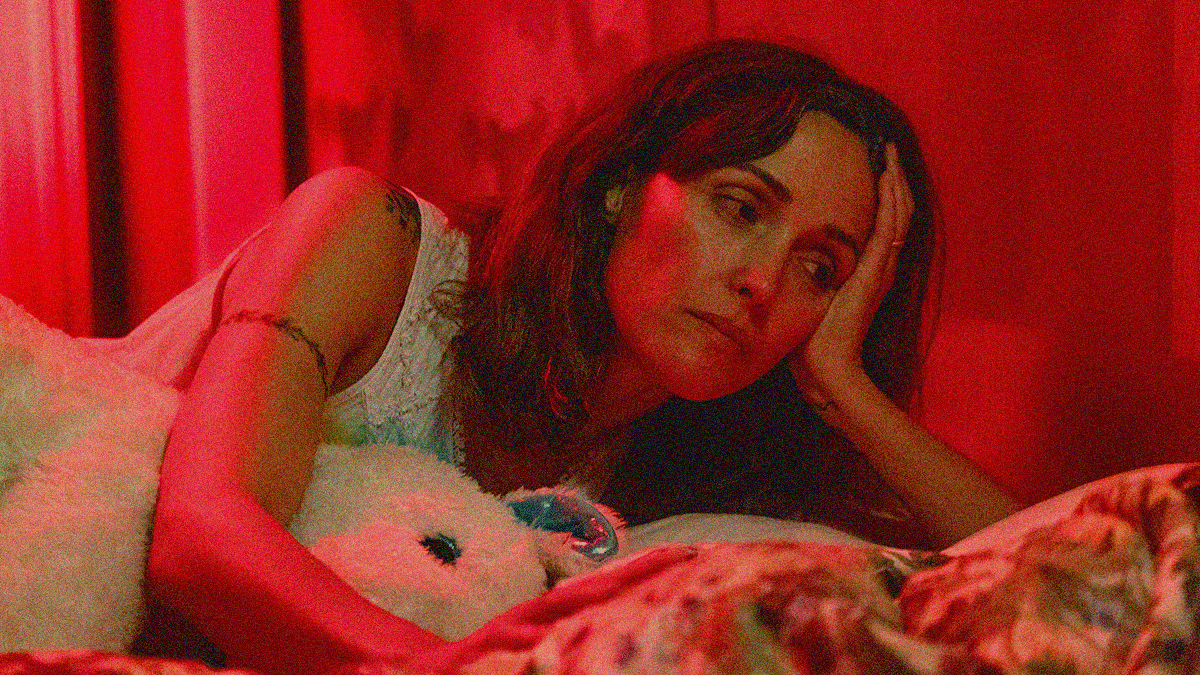 A woman lying on a bed, stuffed animal in one elbow and head resting on the other arm's hand. She looks down at the figure next to her. The whole frame is filtered in red light