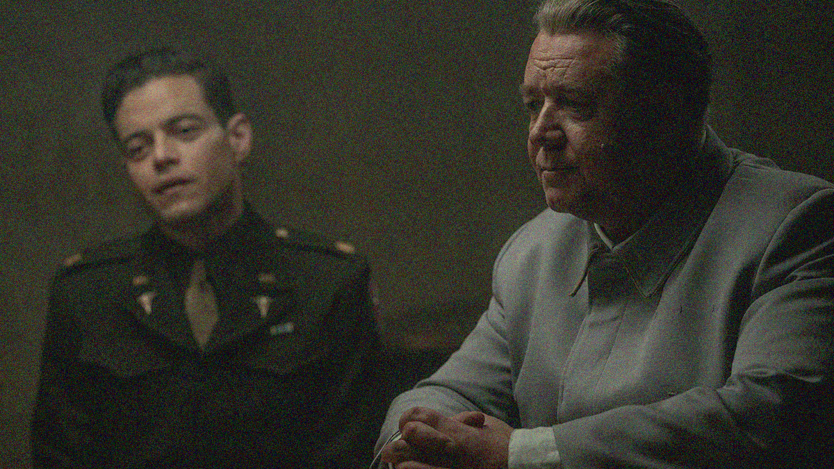 A young man in military uniform sits in the background with head cocked as an older man sits, hands folded, in the foreground.