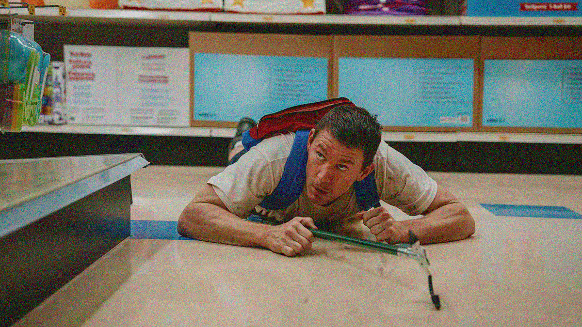 A man with a backpack is belly down on the floor of a department store looking upwards off-screen with a pole grabber in hand.