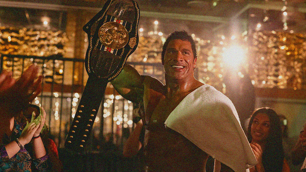 A large muscle-clad man stands amidst a crowd smiling. Towel on left shoulder, championship belt held up by right hand.