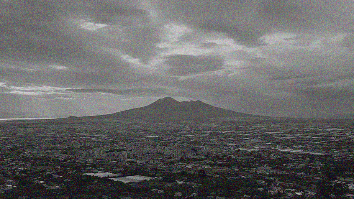Black and white photo of a volcano in the distance beneath dark gray clouds and above a landscape of buildings spanning centuries of time.
