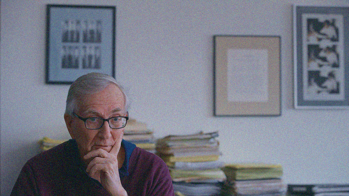 A cropped portrait of a man sitting wherein we see him chest up in the bottom left and a wall of framed photos and piles of papers behind him filling the frame.