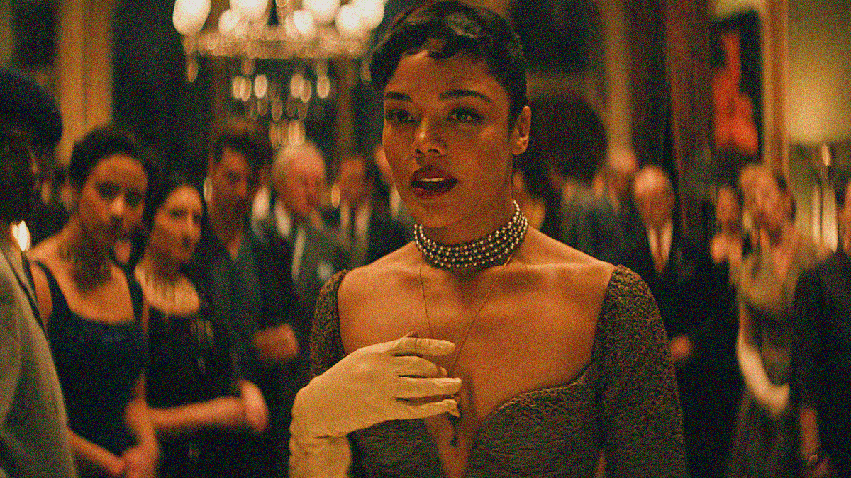 A woman in a low cut dress with necklace wrapped four times around her neck looks off-screen as a room of people behind her do the same.