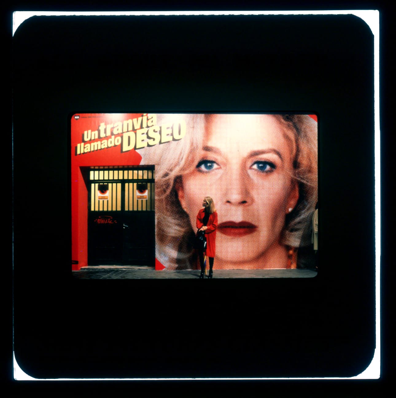 A woman in a red coat is standing against a wall display of another woman's face and the words "Un tranvia llamado deseo." Scale shows her height as being between the photo's chin and nose.