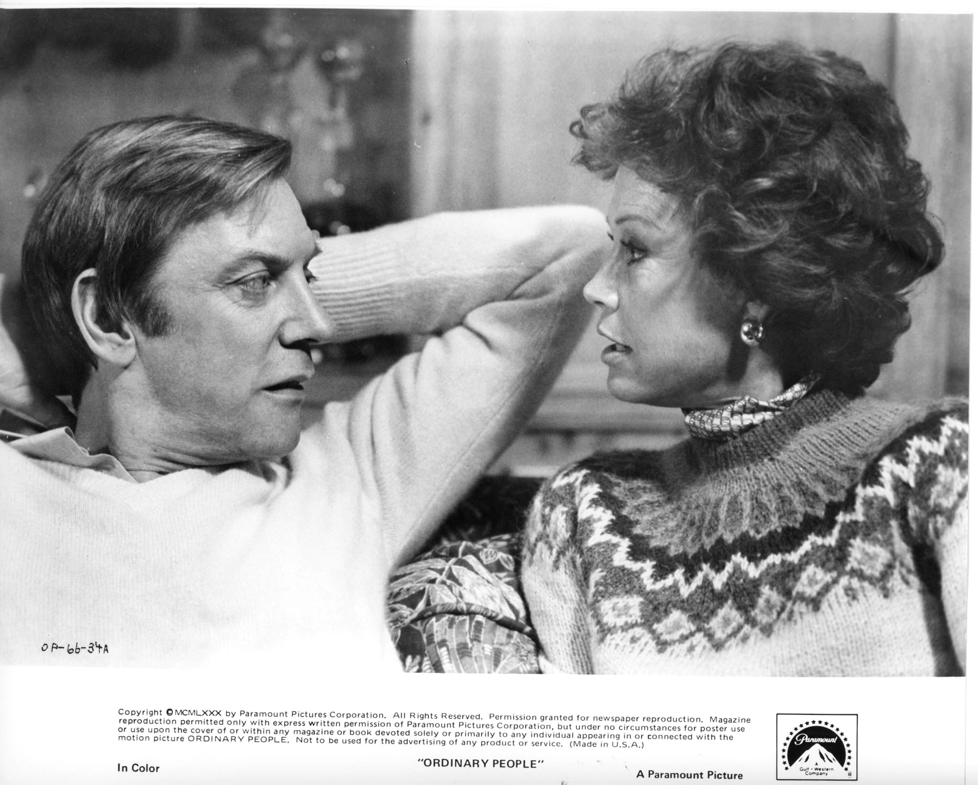 Close-up of a man and woman from the chest up, sitting on a couch in sweaters, looking at each other. 