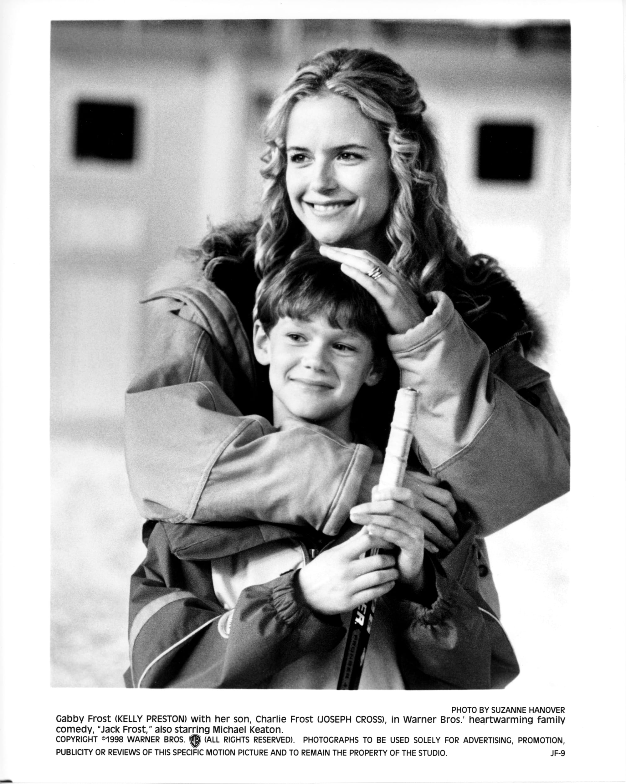 B&W Publicity photo: A woman stands behind her son with one hand on his head and the other around his chest. He holds a hockey stick. Both are smiling while looking off-screen left.