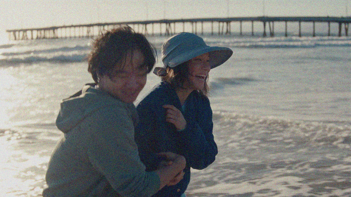 A son in hoodie and mother in sun hat embrace and laugh on the beach with the water and a walking bridge in the background.