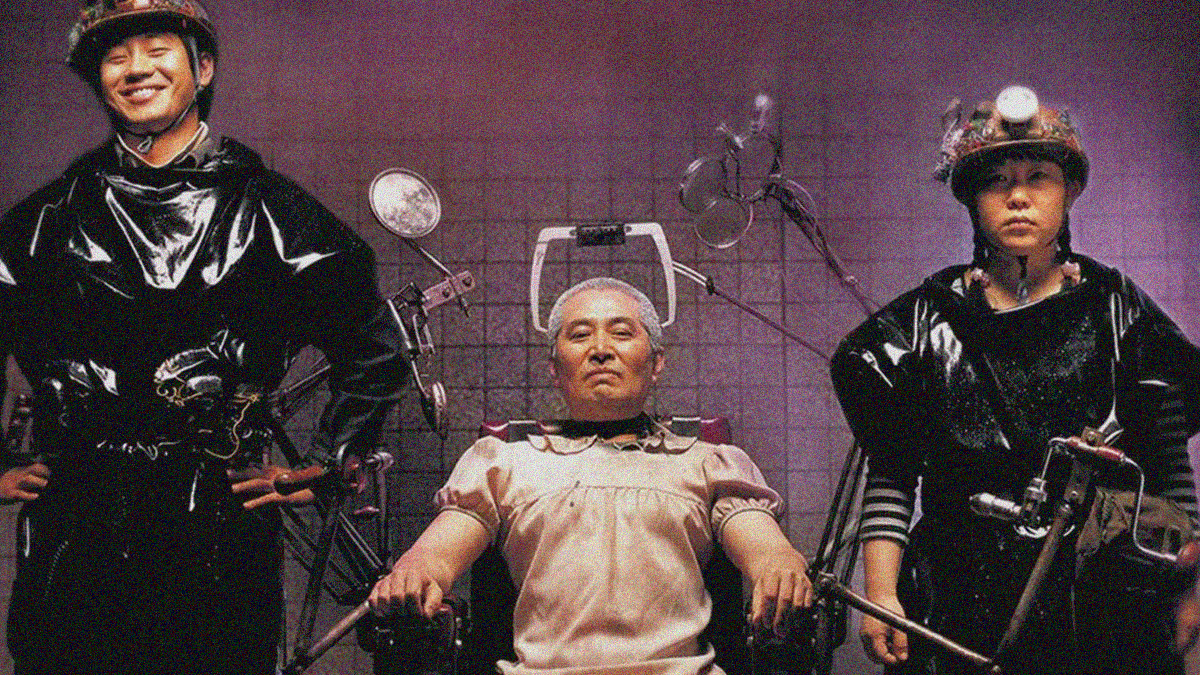A smiling man and stoic woman wearing mechanized hard hats and rubber ponchos flank a bald man in a frilly dress who's strapped to a makeshift operating chair.
